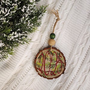 May include: A round wooden Christmas ornament with a natural bark edge. The ornament features a painted green and red design with the word "Joy" in red glitter. It hangs from a twine loop with a green and a wooden bead. The background is a white knitted blanket.
