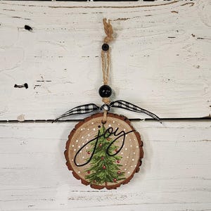 May include: A round, wooden Christmas ornament with a painted green Christmas tree and the word "joy" in black script. The ornament has a twine hanger, a black bead, and a black and white checkered ribbon.