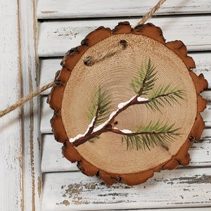 May include: A round wooden slice with a painted pine branch with white snow on it. The slice is brown with a natural bark edge and is hanging from a twine string.