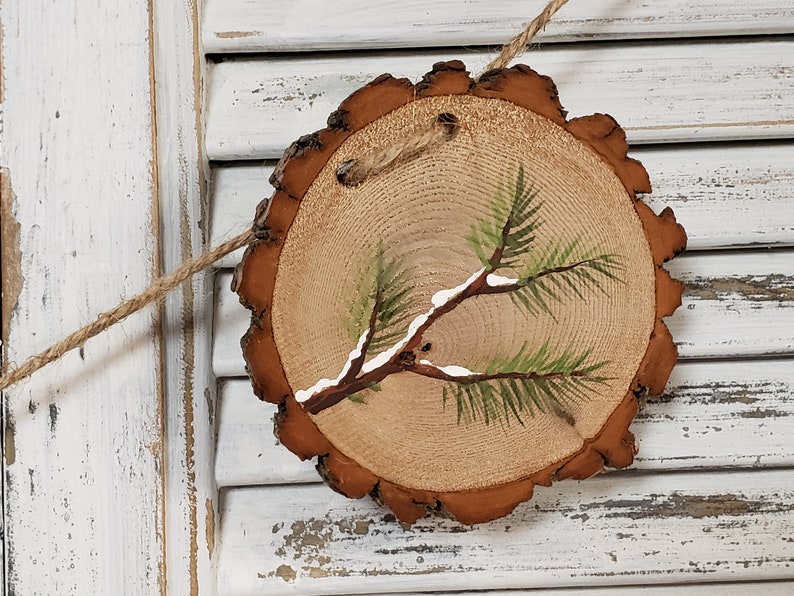 May include: A round wooden slice with a painted pine branch with white snow on it. The slice is brown with a natural bark edge and is hanging from a twine string.