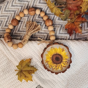 May include: A wooden bead garland with a brown tassel hangs on a white and gray blanket. A wooden round sign with a sunflower and the text "happy fall" is on the blanket.