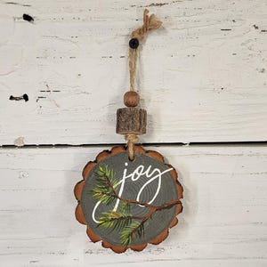 May include: A rustic wooden ornament with a bark edge, painted with a green branch and the word "joy" in white script. It hangs from a twine loop with a wooden bead and a small wooden cylinder.