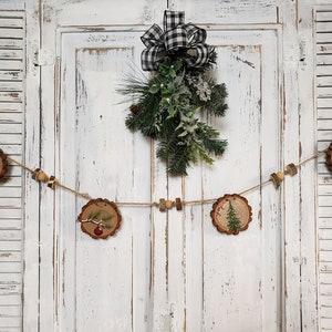 May include: A rustic Christmas garland made of wood slices with painted pine trees and red ornaments. The garland is strung with twine and wooden beads and hangs on a white wooden door with a black and white checkered bow.