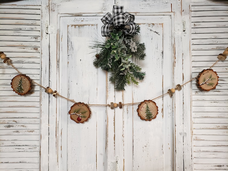 May include: A rustic Christmas garland made of wood slices with painted pine trees and red ornaments. The garland is strung with twine and wooden beads and hangs on a white wooden door with a black and white checkered bow.