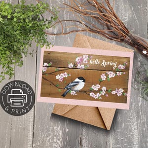 Op de afbeelding: Een printbare kaart met een geschilderd beeld van een vogel die op een tak zit met roze bloesems. De achtergrond is een houtnerfpatroon. De tekst "hello spring" is in wit op de kaart geschreven.