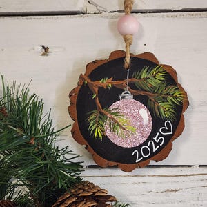 May include: A round, wooden Christmas ornament with a black painted background and a natural wood edge. The ornament features a pink glitter ball, green painted pine needles, and the year "2025" with a heart. It hangs from a jute string with a pink bead.