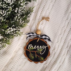 May include: A wooden Christmas ornament with a black center and the word "merry" written in white script. The ornament has a natural wood edge, a black and white checkered bow, and a twine loop for hanging. Green pine branches are painted on the ornament.