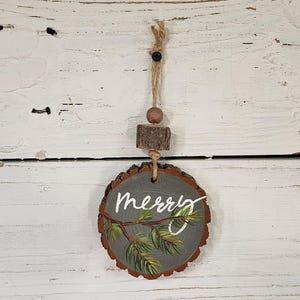 May include: A round, gray wooden ornament with the word "merry" in white script and a painted pine branch. The ornament is attached to a twine loop with a wooden bead and a small wood slice.