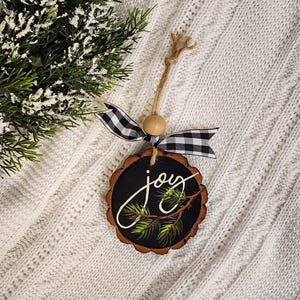 May include: A round, wooden Christmas ornament with a black center and a natural wood edge. The word "joy" is written in white script, with a painted pine branch. It has a twine hanger, a wooden bead, and a black and white checkered ribbon.
