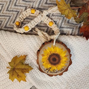 May include: A wooden round sign with a painted sunflower and the word "Autumn" in cursive. The sign is hanging from a white macrame cord with yellow beads. The sign is on a white and gray knitted blanket with fall leaves.
