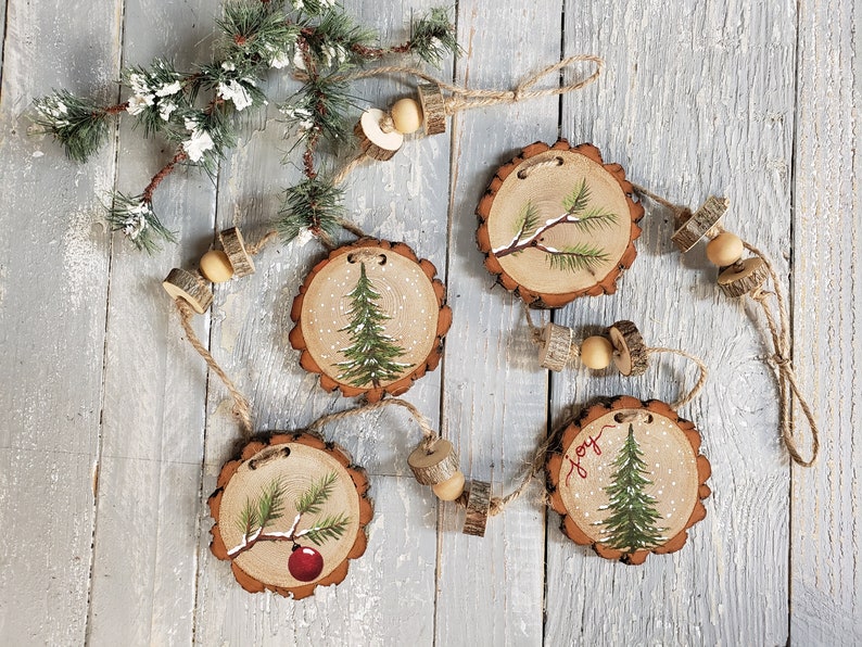 May include: A rustic Christmas garland with four wooden ornaments painted with pine trees and snow. The ornaments are strung together with twine and wooden beads. One ornament has the word "Joy" painted on it.