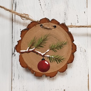 May include: A hand-painted wooden Christmas ornament featuring a pine branch with a red ornament hanging from it. The ornament is painted on a round slice of wood with a rustic brown finish.