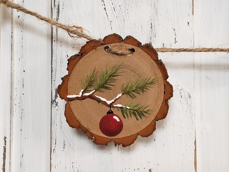 May include: A hand-painted wooden Christmas ornament featuring a pine branch with a red ornament hanging from it. The ornament is painted on a round slice of wood with a rustic brown finish.