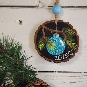 May include: A round, wooden Christmas ornament with a painted design. The ornament features a blue glittery bauble, green pine needles, and the year 2025 with a heart. It hangs from a twine string with a blue bead.