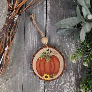 May include: A rustic wooden ornament featuring a hand-painted pumpkin and sunflower design. The orange pumpkin is surrounded by green leaves, with a yellow sunflower at the base. The ornament hangs from a jute string and wooden bead, perfect for fall decor.
