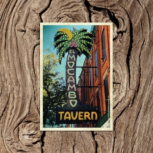 Pode incluir: Um cartão postal vintage com uma placa de néon para "El Mocambo Tavern" com um design de palmeira. A placa está em um prédio de tijolos com uma porta de madeira.