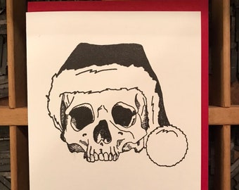 May include: A black and white illustration of a skull wearing a Santa hat. The skull is facing forward with its mouth open. The hat has a white fur trim and a red pom-pom.