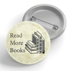 May include: A circular badge featuring the words "Read More Books" and a graphic of stacked books. The badge has a textured, aged appearance and a silver pin. The design is in black and white.