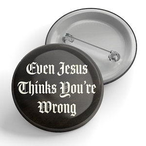 Puede incluir: Un botón negro y blanco con el texto "Even Jesus thinks you're wrong" en una fuente gótica.