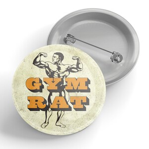 May include: A round, silver-colored pin with a graphic of a muscular man flexing. The text "GYM RAT" is in orange, retro-style lettering. The pin has a distressed, vintage look.