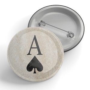 May include: A round button with a white background and a black ace of spades playing card design. The button has a silver metal backing.