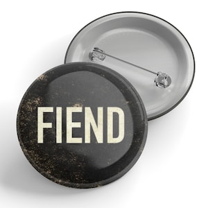 Peut inclure: Un bouton noir et blanc avec le mot "FIEND" en lettres blanches sur un fond noir. Le bouton a un aspect vieilli et délabré.