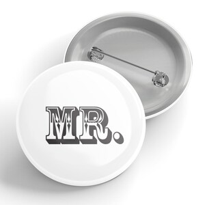 Puede incluir: Botón blanco con un diseño "MR." en blanco y negro. El botón tiene un broche en la parte posterior.