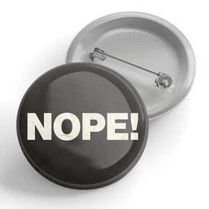 May include: A black button with white "NOPE!" text on it. The button is attached to a silver pin back.