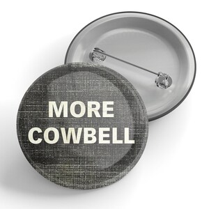 以下が含まれることがあります： 黒と白のテクスチャの背景を持つ丸いボタン。ボタンには「MORE COWBELL」というテキストが白で印刷されています。