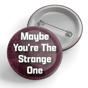 May include: A maroon button with a white background and the text "Maybe You're The Strange One" in white.
