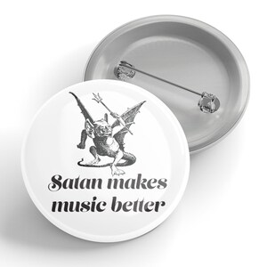 以下が含まれることがあります： 悪魔がトライデントを持っている白黒のイラストが描かれた白いボタン。イラストの下に「Satan makes music better」というテキストが黒で印刷されています。
