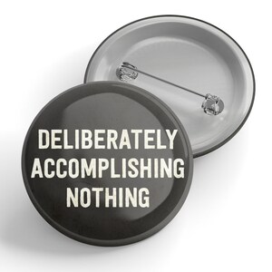 以下が含まれることがあります： 「DELIBERATELY ACCOMPLISHING NOTHING」という白い文字が書かれた黒いボタンです。
