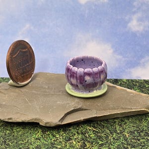 May include: A miniature ceramic planter with a fluted, purple-speckled exterior and a light green saucer base, positioned next to a copper one-cent coin for scale, showcasing a small, rounded vessel design with a glossy, textured finish.