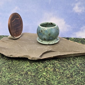 May include: A miniature round ceramic planter with a glossy light blue and green speckled glaze, featuring a matching attached saucer base, positioned next to a United States one-cent coin for scale to demonstrate its small size.
