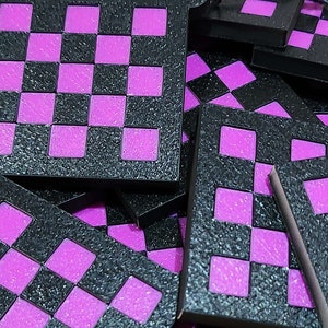 Op de afbeelding: Een stapel zwarte en paarse schaakbordtegels. De tegels zijn gemaakt van een getextureerd materiaal en hebben een schaakbordpatroon met afwisselend zwarte en paarse vierkanten.