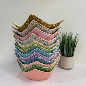 Peut inclure: Une pile verticale de bols en tissu carrés présentant divers motifs, notamment des imprimés floraux, des carreaux vichy et des pois dans des couleurs comme le jaune moutarde, le rose, le vert et le bleu, avec une petite plante verte à côté.