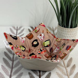 May include: A beige fabric bowl with a sushi pattern on the inside. The bowl is square-shaped with a folded design. The sushi pattern includes images of sushi rolls, noodles, and other Japanese food items. The bowl is sitting on a patterned surface.