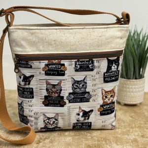 Peut inclure: Un sac bandoulière avec une sangle marron clair et un panneau supérieur crème. Le corps principal présente un tissu clair imprimé de portraits de chats façon photo d'identité judiciaire, avec des mentions "Wanted" et des délits humoristiques.