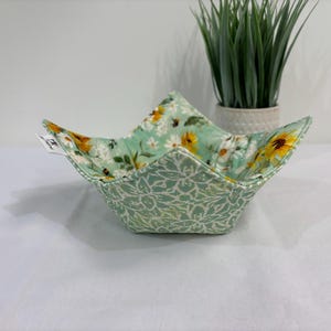 Peut inclure: Un bol en tissu avec un intérieur vert menthe clair orné de tournesols jaunes, de marguerites blanches et de petites abeilles. L'extérieur est en tissu vert menthe clair avec un motif floral blanc. Une petite étiquette blanche est fixée sur le côté.