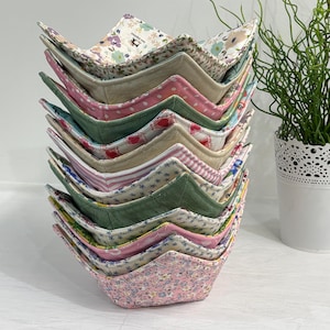 May include: A vertical stack of ten fabric bowl cozies featuring various patterns including floral prints, polka dots, and stripes in shades of pink, green, blue, and cream. Each cosy has a square base and four folded corners, creating a star-like shape.