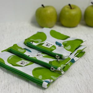 May include: Three zippered pouches with a green apple pattern on a white background. The pouches have green zippers and trim. Three green apples are in the background.