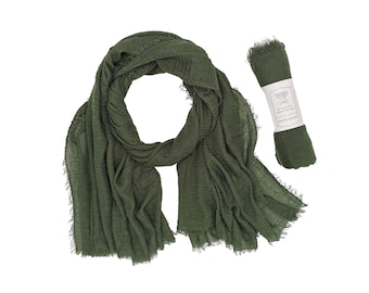 May include: Olive green scarf with fringed edges, draped and rolled. The scarf is made of a lightweight fabric. A label on the rolled scarf reads "MADIRA Natural Fine Silk".