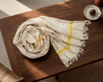 May include: A rolled, off-white linen scarf with yellow stripes and fringed edges rests on a dark wood table. The scarf is next to a small, decorated plate with a brown object.