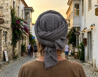 May include: A person wearing a gray head wrap and a brown shirt, viewed from behind, stands on a cobblestone street. The background features stone buildings, flowering plants, and people walking in the distance. The sky is a warm orange.