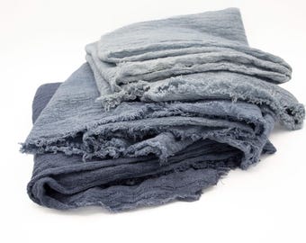 May include: A stack of several blue-toned fabric cloths. The cloths are layered, with varying shades of blue, from light to dark. The edges of the fabric have a frayed, textured appearance, adding to the rustic aesthetic.