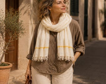 May include: A woman wearing a cream-colored scarf with yellow stripes and fringe, paired with a brown button-down shirt and light-colored pants. She is carrying a woven tote bag with leather handles. The scene is set on a sunlit street.