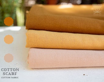 May include: Three folded cotton scarves stacked horizontally, featuring solid colors in deep mustard yellow, golden yellow, and soft pale pink. The text "COTTON SCARF" and "COTTON FABRIC" appears in the bottom left, with a logo in the top right.