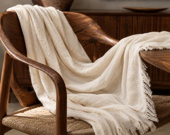 May include: A cream-colored throw blanket with fringed edges draped over a wooden chair. The chair has a woven seat and curved wooden arms. The blanket's texture is visible, suggesting a soft, cozy material. The scene evokes a sense of comfort and relaxation.