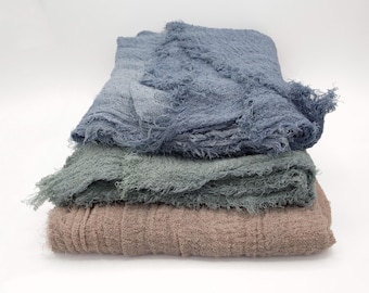 May include: Three folded, textured blankets in shades of blue, green, and tan. The blankets have a frayed edge, and are stacked on top of each other against a white background. The top blanket is a dusty blue color.