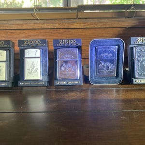 May include: Five Zippo metal lighters displayed in individual packaging. Four lighters are housed in black plastic stands labelled Zippo U.S.A. Lighter and Lifetime Guarantee, while one is in a silver-rimmed case. Each features unique engravings.